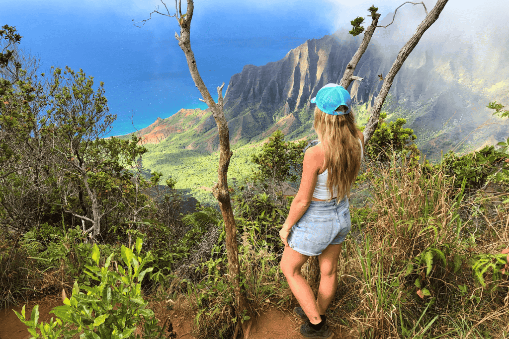 Kate, in a blue hat and denim shorts, stands on a lush cliffside, gazing out at the dramatic ridges and turquoise waters of Kauai’s Nā Pali Coast.