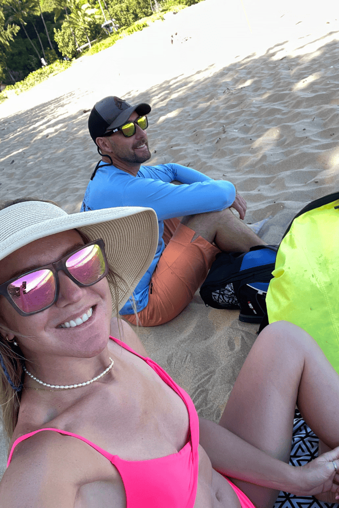 Kate and her companion sit in the sand at Tunnels Beach, smiling in sunglasses and beachwear with tropical greenery in the background.
