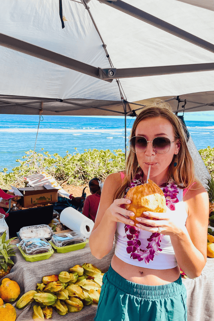 Kate sips from a fresh coconut under a market tent by the ocean, wearing a purple flower lei and sunglasses, with tropical fruits and local treats displayed on the table beside her.