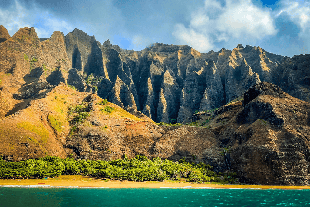 A stunning view of the Nā Pali Coast in Kauai shows jagged emerald and rust-colored cliffs rising sharply behind a narrow stretch of golden beach and turquoise ocean.