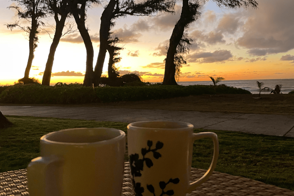 Two coffee mugs sit on a woven table at sunrise, with silhouetted trees, a golden sky, and the ocean in the background on a peaceful Kauai morning.