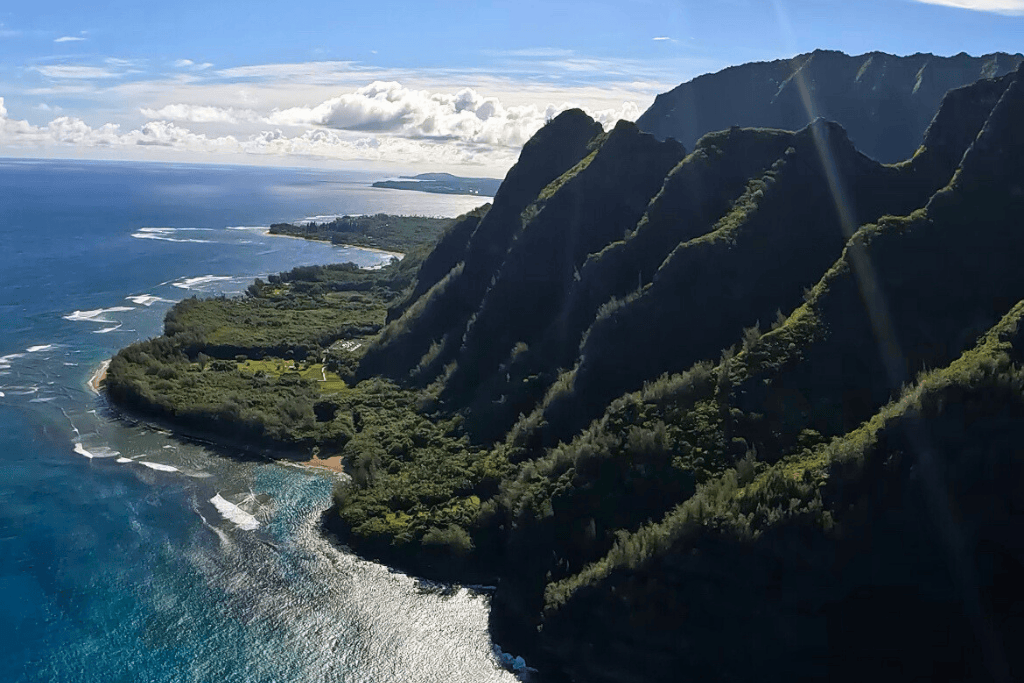 Aerial view of Kauai’s lush, dramatic north shore with ridged emerald cliffs meeting the deep blue Pacific Ocean under a bright sunny sky.