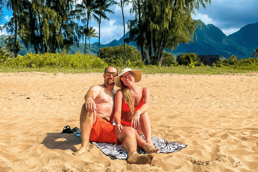 Kate and her companion relax on a beach towel in the golden sand, surrounded by lush greenery and towering mountains under a partly cloudy Kauai sky.
