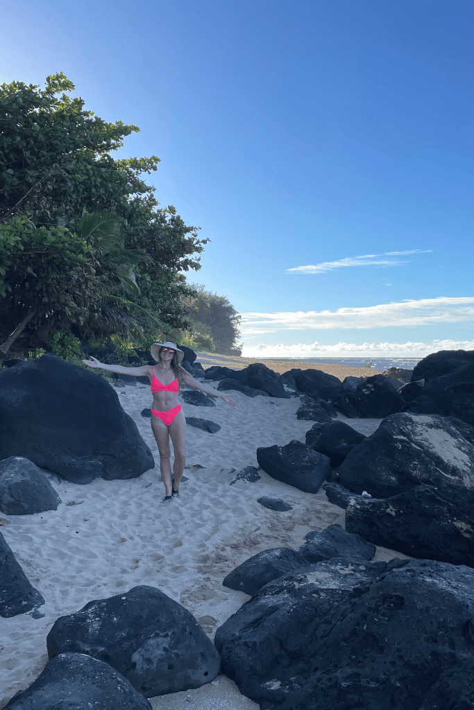 Kate stands on the white sand between black lava rocks at Tunnels Beach, wearing a bright pink bikini and sunhat under a clear morning sky.