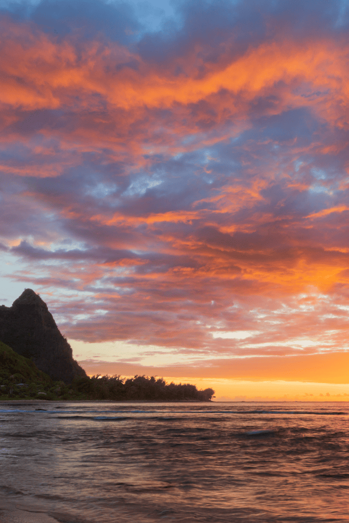 A dramatic sunset at Hanalei Beach lights up the sky in vivid orange and purple hues, with ocean waves gently rolling in beside the silhouette of Kauai’s lush cliffs.