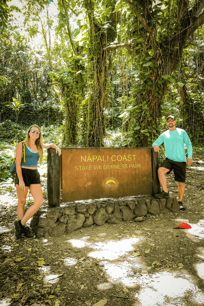 Kate and her companion pose beside the Nā Pali Coast State Wilderness Park sign surrounded by dense jungle foliage within Hā‘ena State Park.