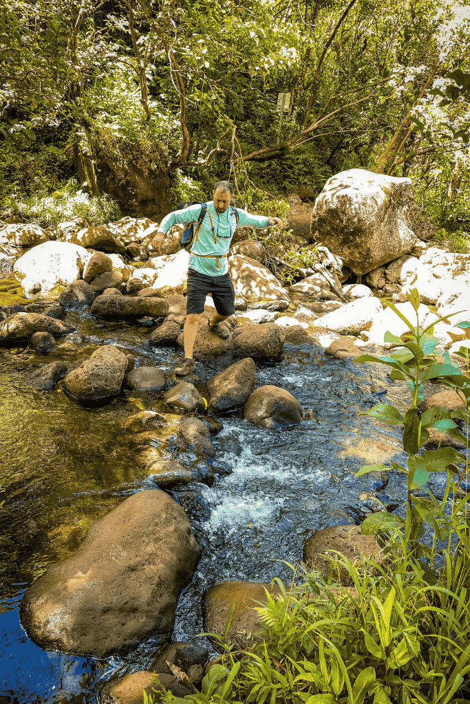 Kate’s companion carefully crosses a stream on large rocks surrounded by dense jungle while hiking to Hanakapiʻai Falls.