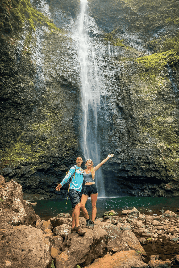 Kate and her companion pose joyfully on boulders at the base of Hanakapiʻai Falls, a towering waterfall plunging into a clear pool surrounded by steep cliffs.