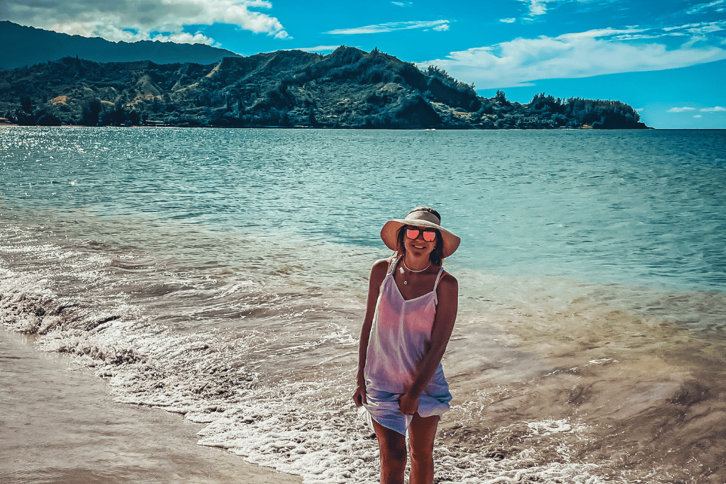 Kate stands at the edge of the shoreline at Hanalei Bay in a sunhat and sunglasses, smiling with lush green cliffs and sparkling blue water behind her in Kauai.