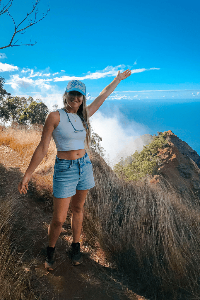 Smiling Kate in a blue “Aloha” hat and hiking boots stands on a trail, raising her arm toward the ocean view and lush ridgeline partially veiled in mist behind her.