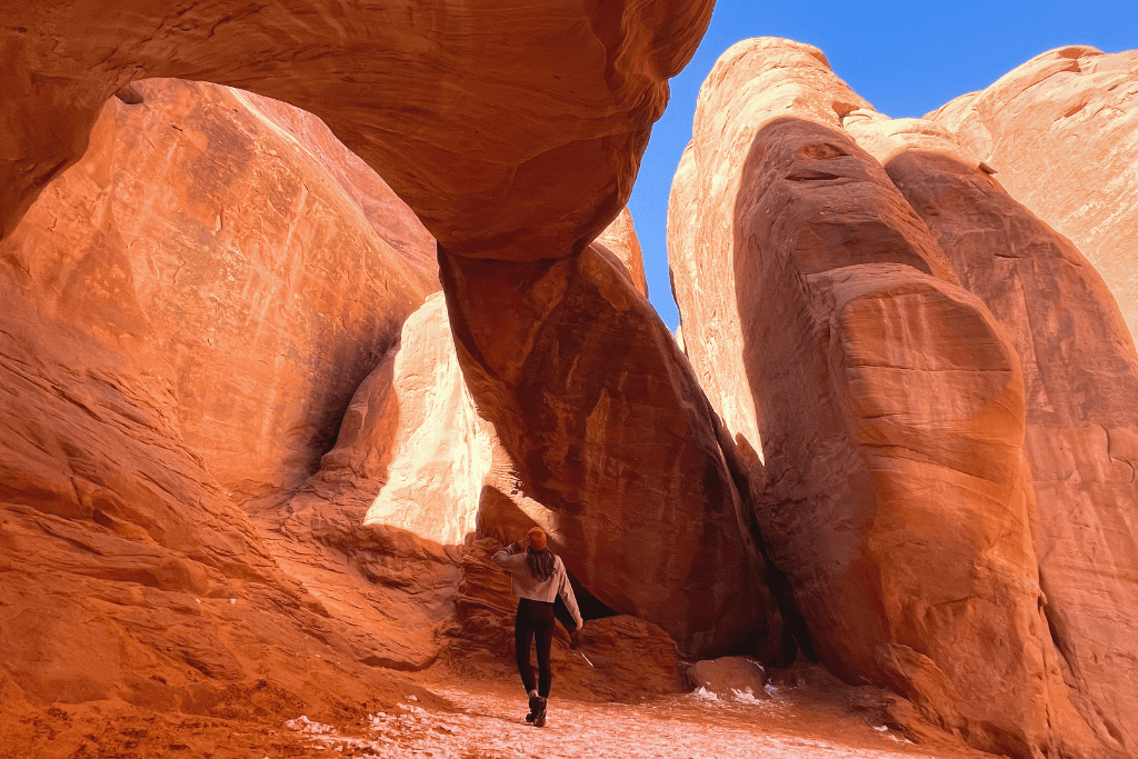 Kate walks through a narrow sandstone canyon beneath a suspended rock fin in Arches National Park, surrounded by glowing red walls and bright blue sky.