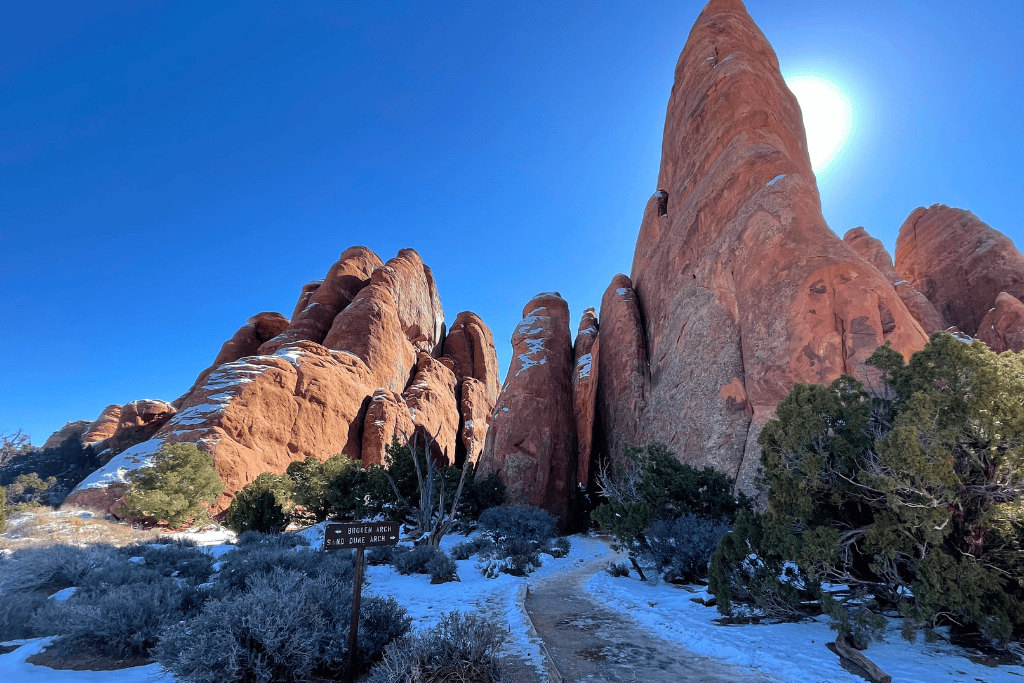 Snow-dusted trail leads toward towering red rock fins in Arches National Park, with a trail sign pointing to Broken Arch and Sand Dune Arch under a bright winter sun.