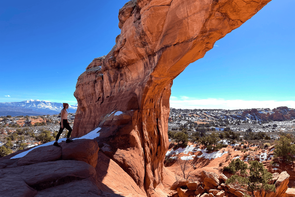 Kate stands beside a tall, slender sandstone arch in Arches National Park, overlooking a snowy desert valley with distant mountains under a clear blue sky.