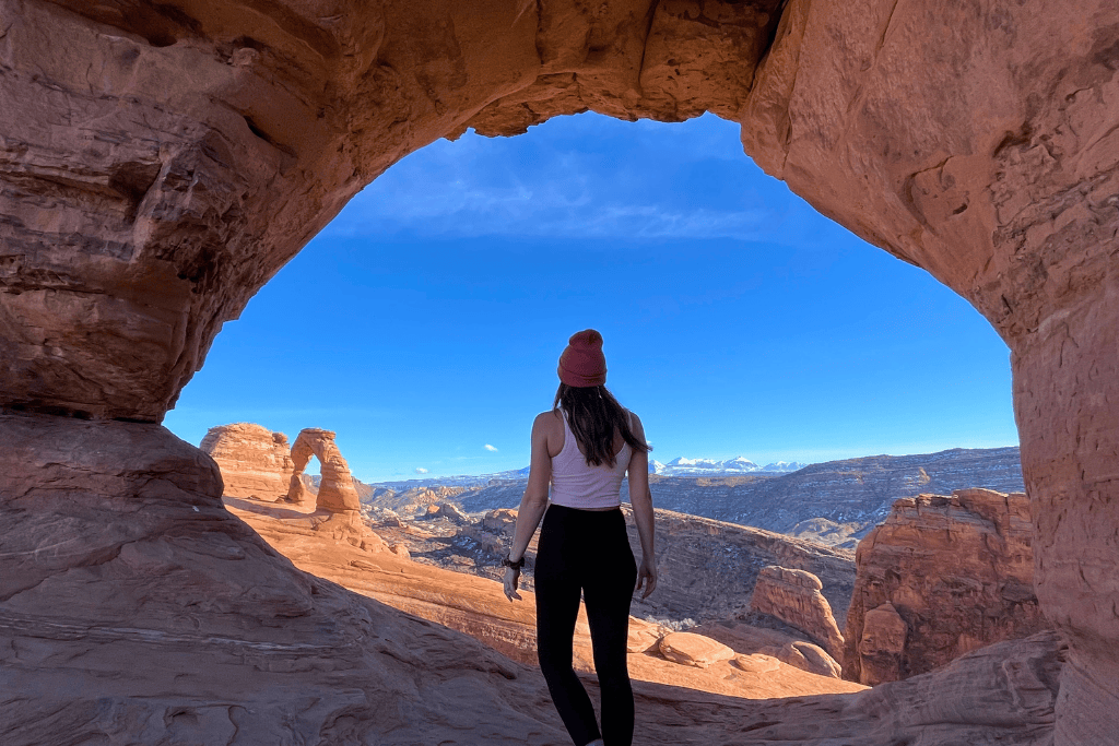 Kate stands framed by a natural sandstone arch, looking out toward Delicate Arch and the snow-capped La Sal Mountains in the distance at Arches National Park.