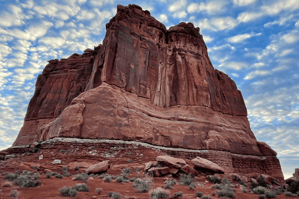 A towering red sandstone formation rises dramatically from the desert floor under a textured, cloud-filled sky in Arches National Park.