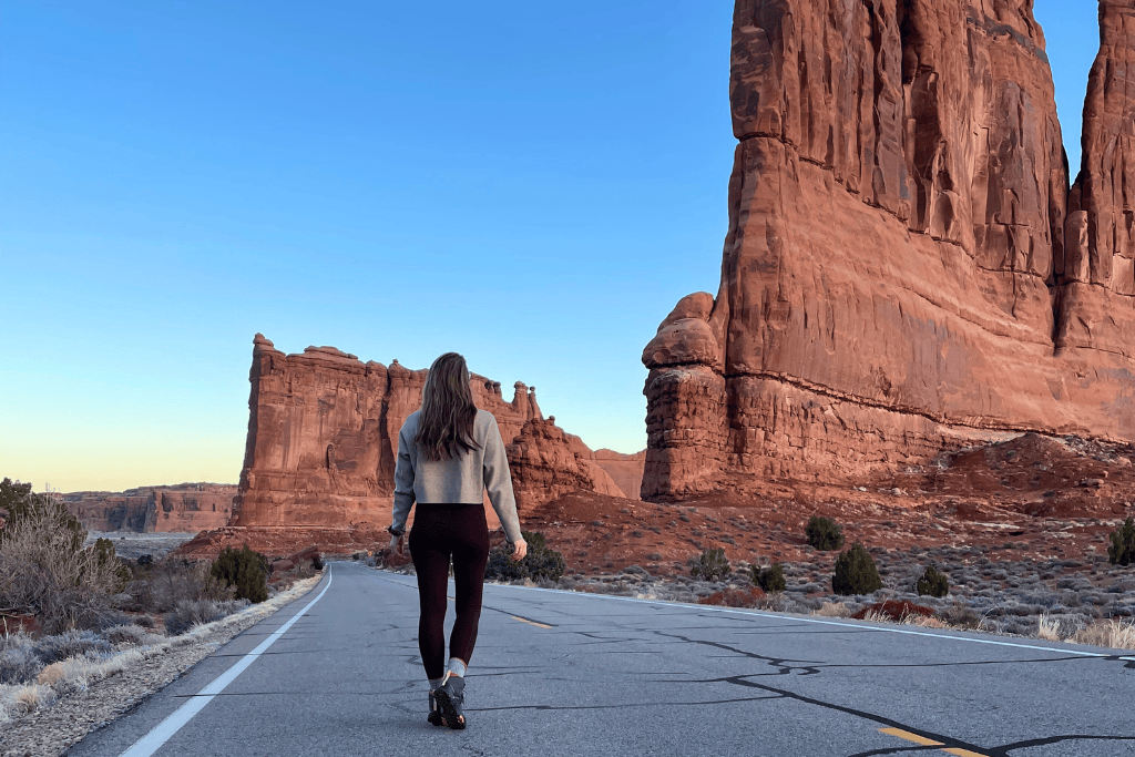 Kate walks down an empty road through Arches National Park, flanked by towering red rock formations in the soft light of early morning or dusk.
