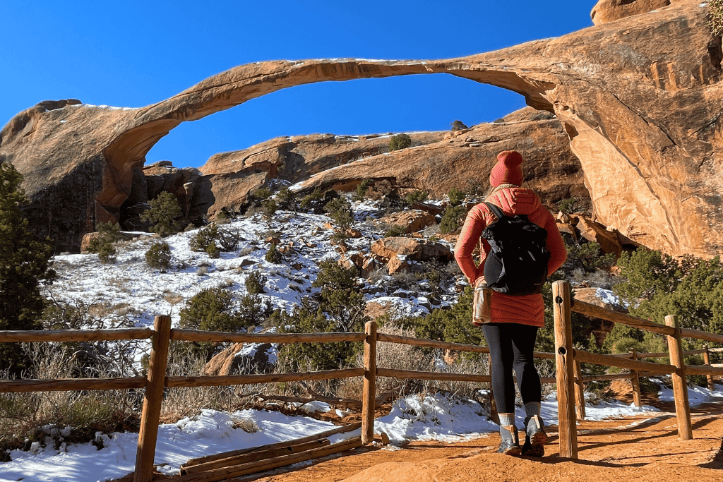 Kate stands at a wooden fence, looking up at the long, slender span of Landscape Arch with snow-covered ground and red rock cliffs in Arches National Park.