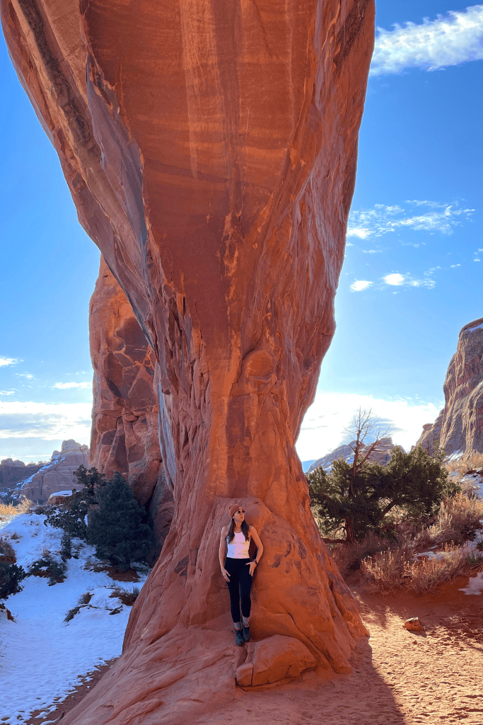 Kate leans against the base of a towering, narrow sandstone arch in Arches National Park, surrounded by snowy patches and desert vegetation under a clear sky.