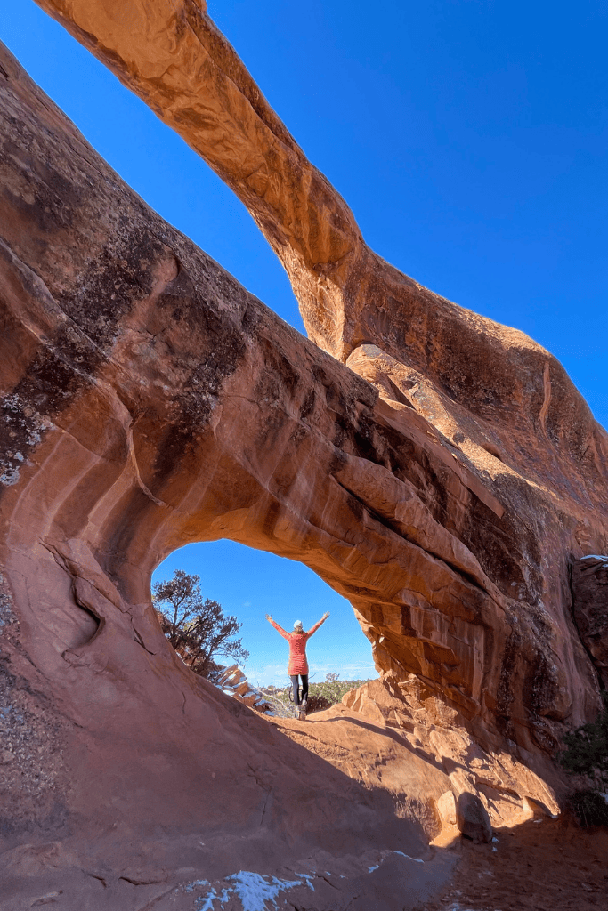 Kate stands with arms raised beneath the Double O Arch in Arches National Park.