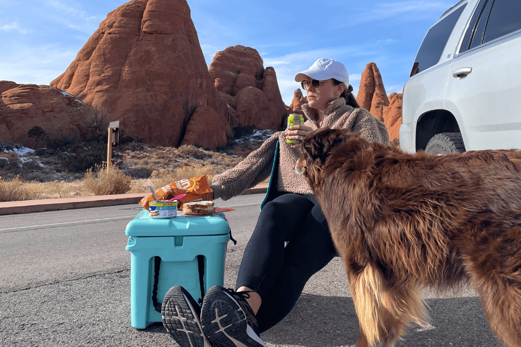 Kate relaxes on the pavement beside her car in Arches National Park, sipping a drink with snacks on a cooler and her brown dog standing nearby.