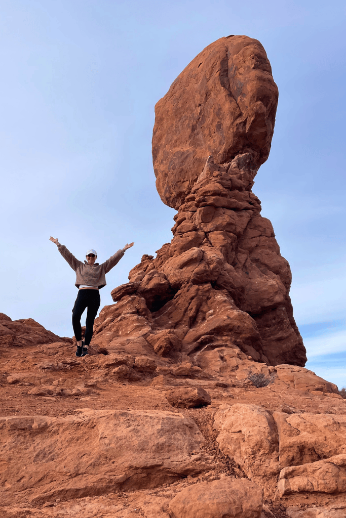 Kate poses with arms raised in front of Balanced Rock, a massive sandstone boulder perched atop a narrow pedestal in Arches National Park.