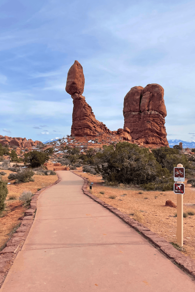A paved trail leads through the desert toward the towering, iconic Balanced Rock formation in Arches National Park, with signs indicating no bikes or pets allowed.