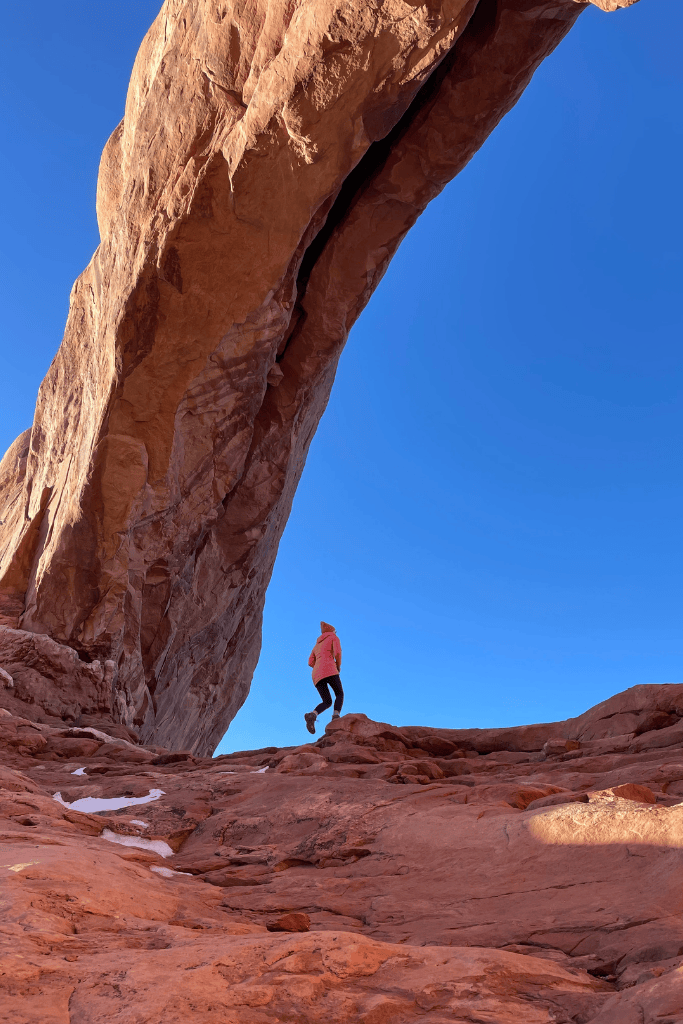 Kate hikes up a rocky slope beneath a towering, narrow rock arch in Arches National Park, with deep blue sky stretching overhead.