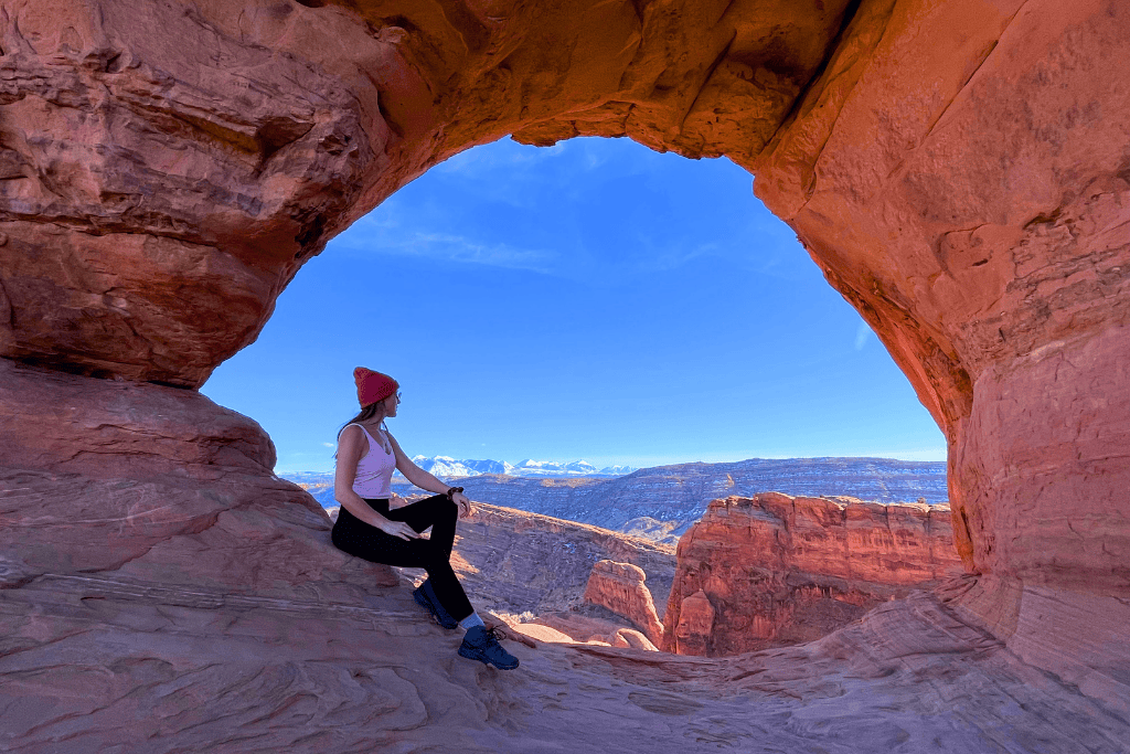 Kate sits inside a natural rock window in Arches National Park, gazing out at the red rock landscape and snow-capped La Sal Mountains under a clear blue sky.