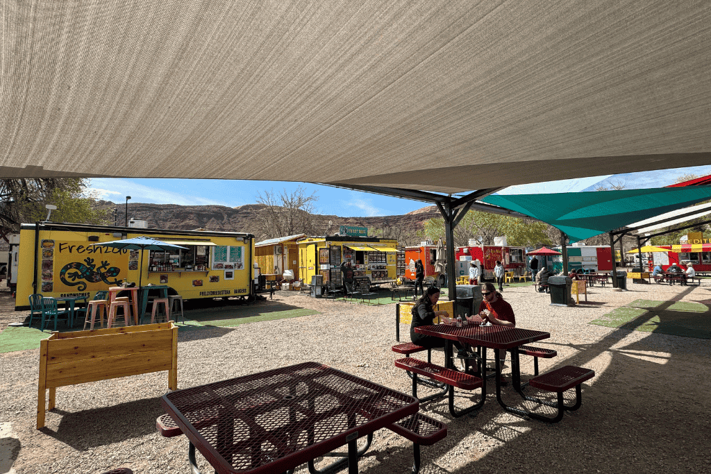 Outdoor food truck park in Moab with colorful vendors, shaded picnic tables, and diners enjoying meals under sun sails with red rock cliffs in the background.