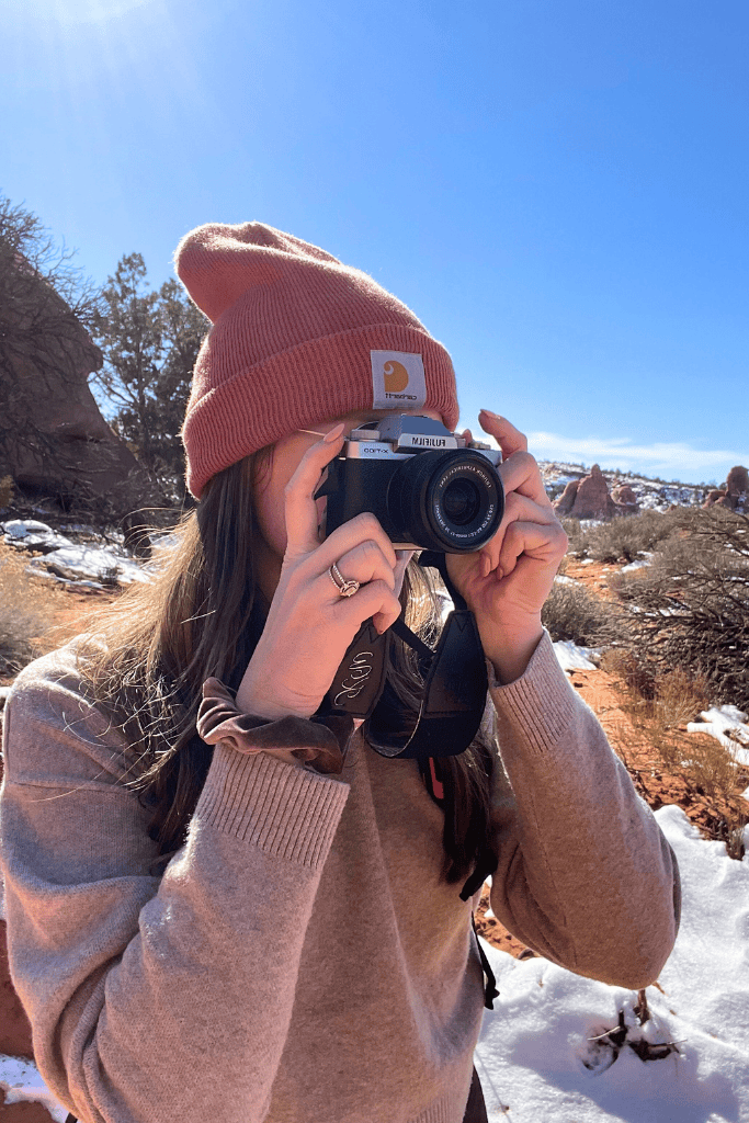 Kate holds a Fujifilm camera to her eye, mid-shot, while exploring a snowy desert trail in Arches National Park.