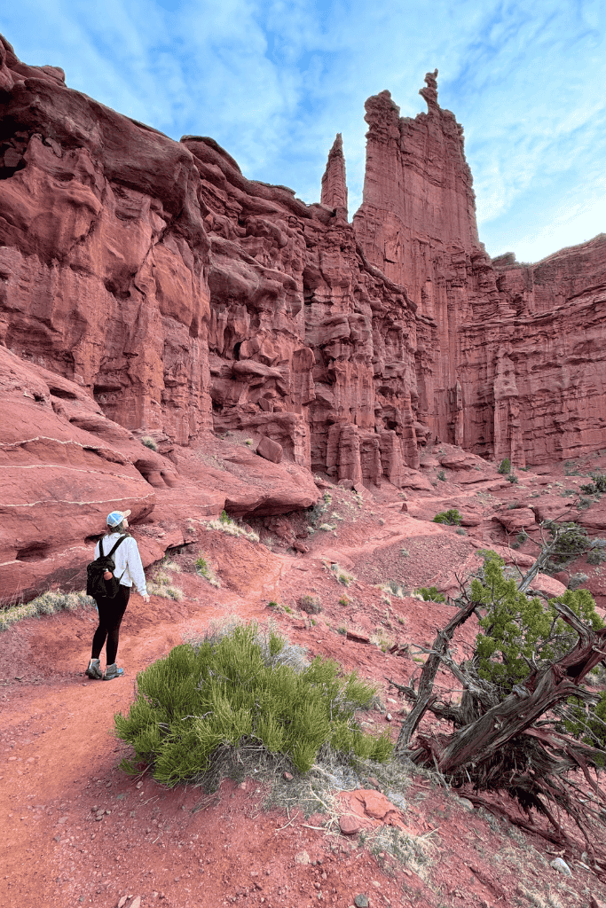 Kate hikes along a red dirt trail through towering, jagged sandstone spires and cliffs in Fisher Towers near Moab, Utah, under a partly cloudy sky.