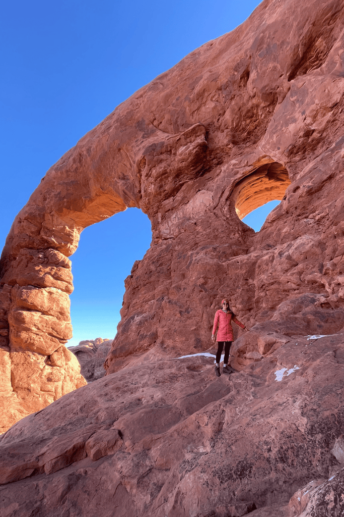 Kate stands on a rocky ledge below two striking sandstone arches carved into a massive red cliff in Arches National Park.