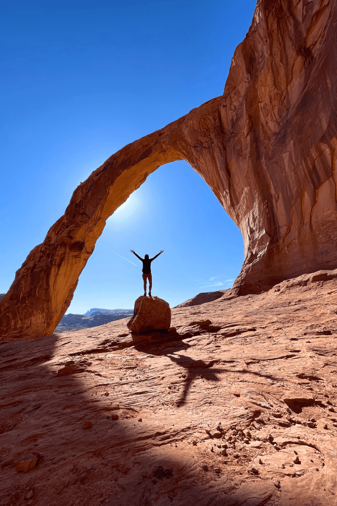 Kate stands triumphantly on a boulder beneath Corona Arch, arms raised as sunlight streams through the massive sandstone curve in the Utah desert.