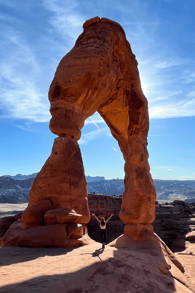 Kate stands with arms raised beneath Delicate Arch in Arches National Park, with sunlight streaming through the iconic sandstone formation against a clear desert sky.