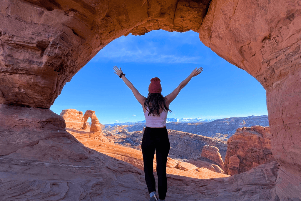 Kate stands with arms raised inside a rock window in Arches National Park, looking out at Delicate Arch and the snow-capped La Sal Mountains in the distance.