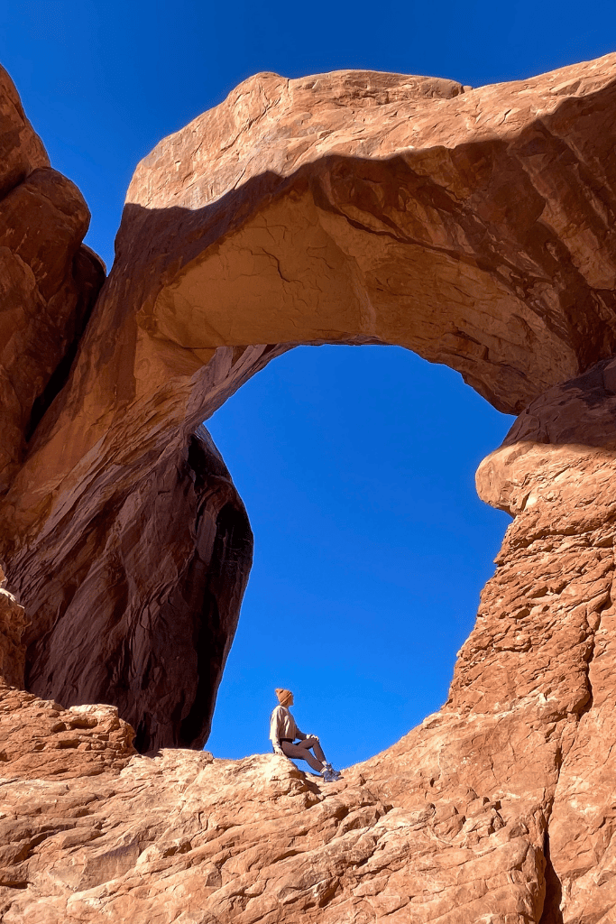Kate sits on a sunlit rock ledge beneath a towering sandstone arch, looking up at the red formation against a brilliant blue sky in Arches National Park.