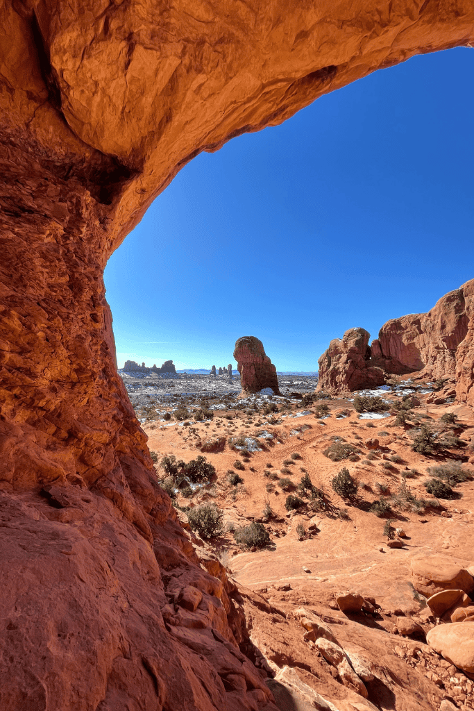 View from beneath a red sandstone arch in Arches National Park, looking out over a snowy desert landscape dotted with rock formations under a cloudless blue sky.