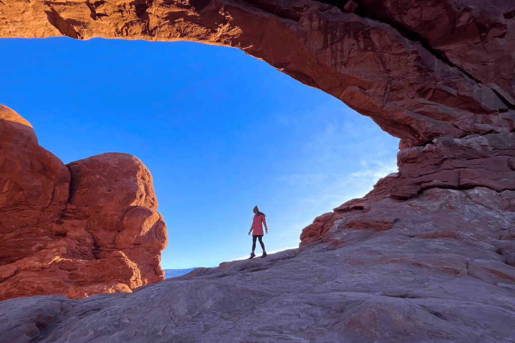 Kate stands beneath a massive sandstone arch in Arches National Park, surrounded by red rock formations and a brilliant blue sky.