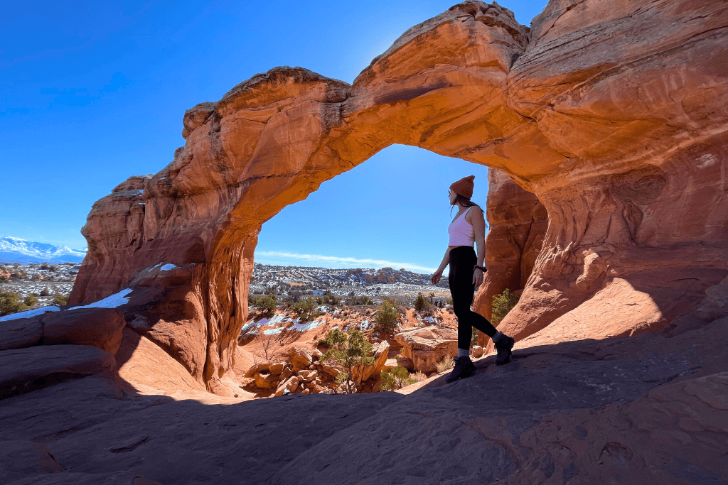 Kate stands beneath a striking red sandstone arch, gazing out over the snowy desert landscape under a bright blue sky.