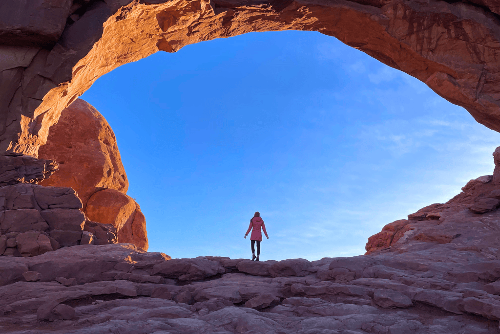 Kate stands silhouetted beneath a massive arch in Arches National Park, framed by glowing red rock and a vivid blue sky.