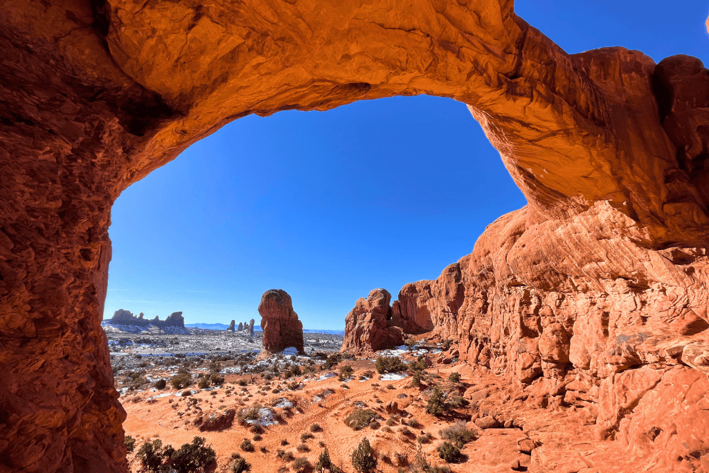 A sweeping desert landscape dotted with red rock formations is framed by the curve of a sandstone arch under a brilliant blue sky in Arches National Park.