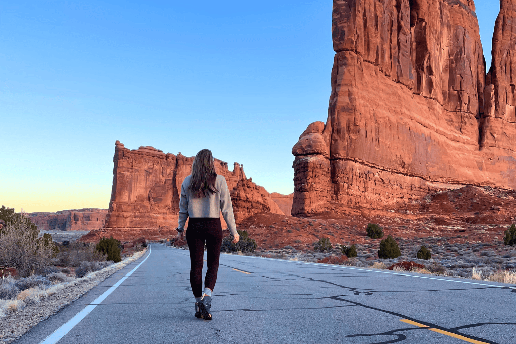 Kate walks alone down an empty road in Arches National Park, surrounded by towering red sandstone cliffs glowing in the soft light of sunrise or sunset.