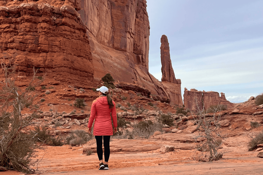 Walking along the red dirt trail, Kate approaches the dramatic monoliths of Park Avenue, surrounded by rugged desert terrain.