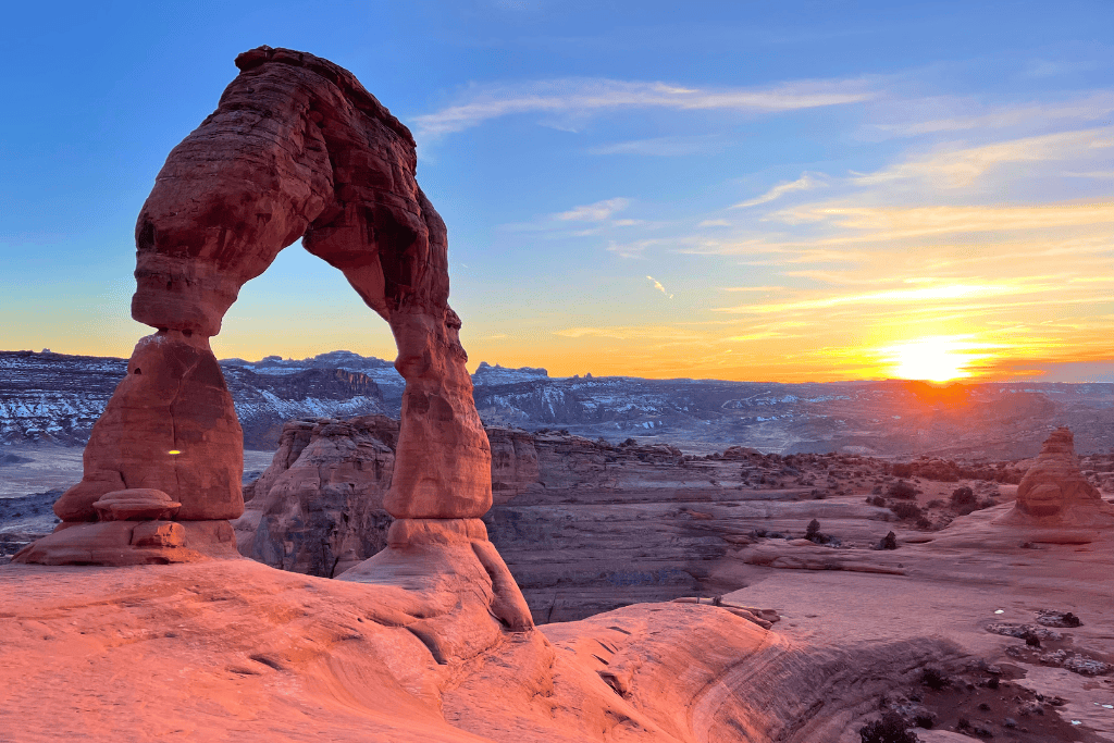 The iconic Delicate Arch glows in the warm light of sunset, standing tall against a backdrop of snowy mesas and a colorful desert sky.