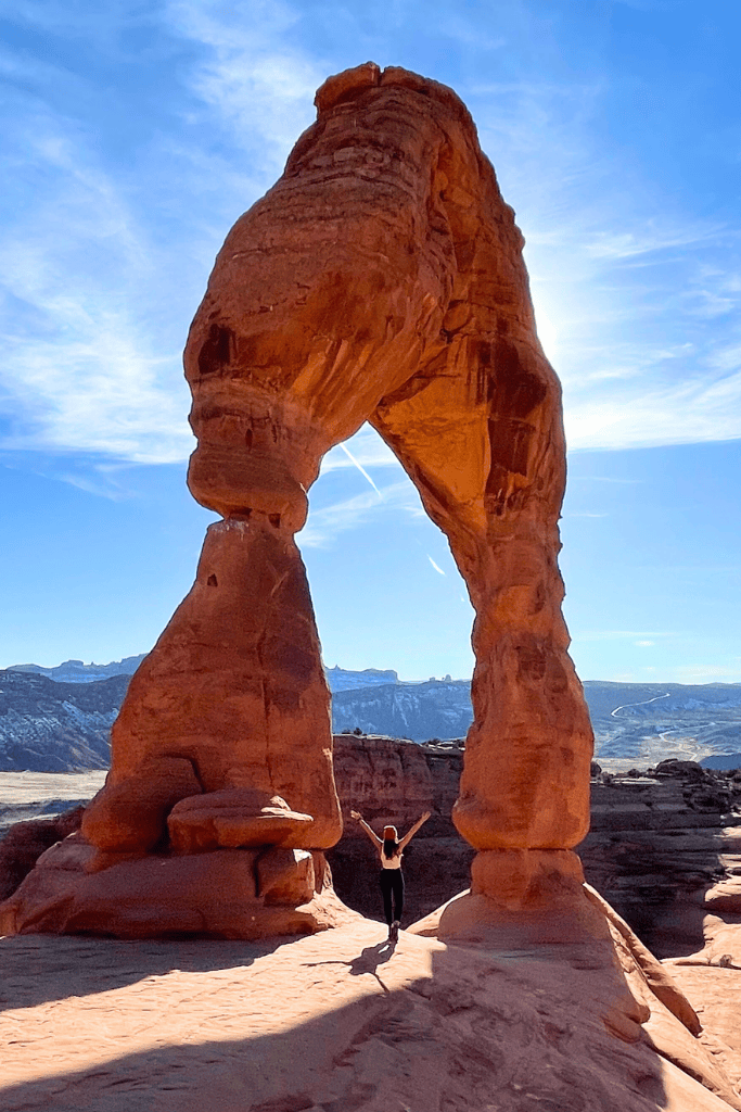 Kate raises her arms in triumph beneath Delicate Arch, perfectly silhouetted against a bright midday sun in Arches National Park.