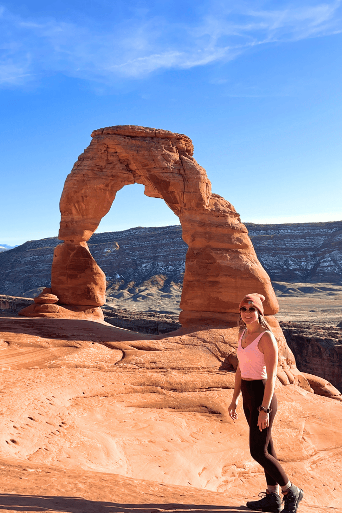 Kate smiles in front of Delicate Arch, standing on sunlit red rock with the iconic formation and La Sal Mountains in the background.