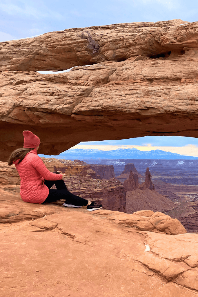 Kate sits beneath Mesa Arch, looking out over a dramatic canyon landscape framed by the sandstone arch and distant snow-capped peaks.
