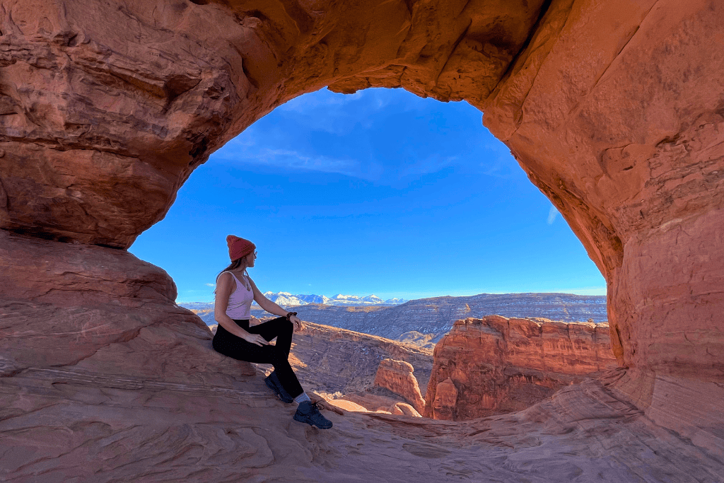Kate sits beneath a red sandstone arch, gazing out at the sweeping desert landscape and snow-capped La Sal Mountains in the distance.