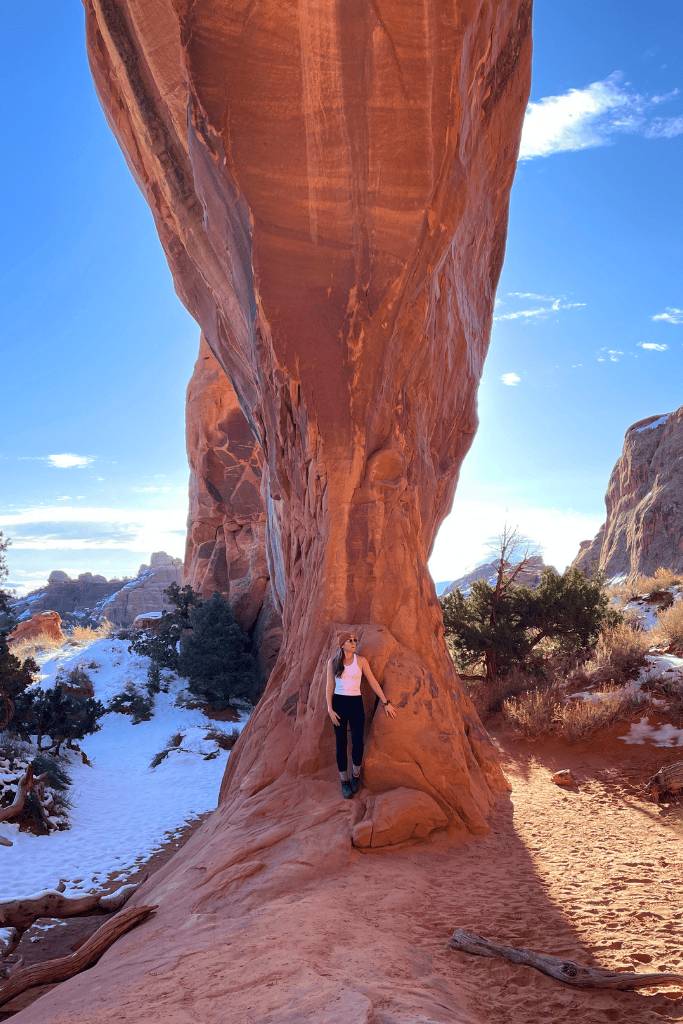 Kate stands at the base of the towering, slender rock formation known as Landscape Arch in Arches National Park, its massive span rising dramatically above her against a clear winter sky.