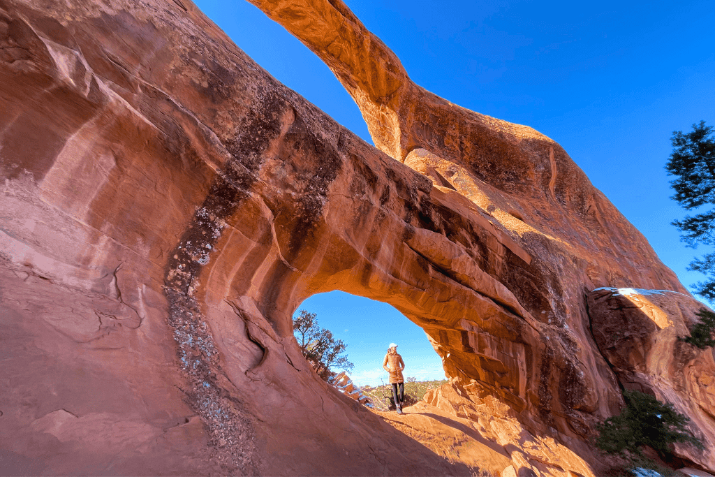 Kate stands with arms raised beneath the towering Double Arch, dwarfed by the massive sandstone spans reaching into the clear blue sky.